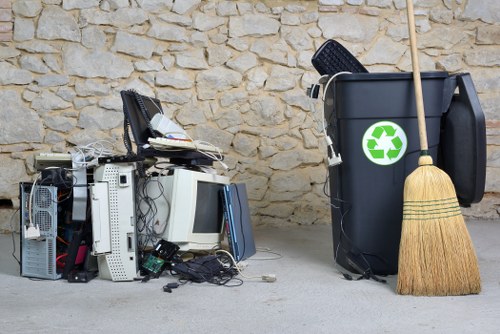 The image shows a collection of discarded electronic waste and household rubbish placed on a concrete surface in front of a natural stone wall. On the left, various broken or obsolete computer equipment and components, including a computer tower, monitors, keyboards, and scattered cables, are piled together with some parts lying on the ground. To the right, there is a large grey rubbish bin featuring a green recycling symbol, with a black lid ajar and a slim wooden broom with a straw brush leaning against it. A black plastic dustpan is resting on top of the bin. The scene suggests a rubbish removal or disposal situation, potentially related to office or domestic clearance near Kentish Town, with the visible objects illustrating typical electronic and general waste that would be handled by professional rubbish removal services such as those offered by Office Clearance Kentish Town.