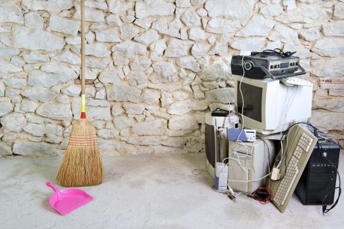 The image depicts a collection of discarded electronic equipment and household items placed on a tiled floor against a stone wall. Included are an old computer monitor, a tower desktop computer, an older style beige telephone, and a beige keyboard. There are also various tangled cables and wires among the electronic devices. To the left of the electronics, a pink plastic dustpan with a curved handle lies on the floor, alongside a traditional straw broom with a red and yellow handle leaning against the wall. The scene appears to be in a residential or storage area, indicative of rubbish awaiting removal or disposal. This setup aligns with a rubbish removal service such as those offered by Office Clearance Kentish Town, specialising in clearing clutter and electronic waste in the local area, possibly near Kentish Town or postcode NW5.