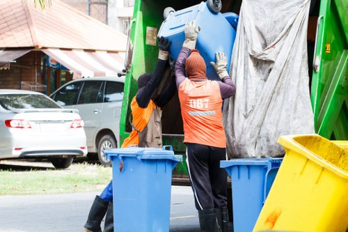 Two waste collection workers wearing high-visibility orange vests and gloves are lifting a large blue plastic bin into a green rubbish collection vehicle parked on a residential street. The workers stand on the pavement, surrounded by additional blue and yellow bins, with a suburban background that includes parked cars, a building with a striped awning, and trees. The vehicle's side panel is partially visible, and the overall scene depicts a routine rubbish removal operation typical of local services in Kentish Town or nearby areas. The environment appears bright and clear, with natural daylight illuminating the scene, emphasizing the practical and professional nature of the waste collection activity carried out by Office Clearance Kentish Town.
