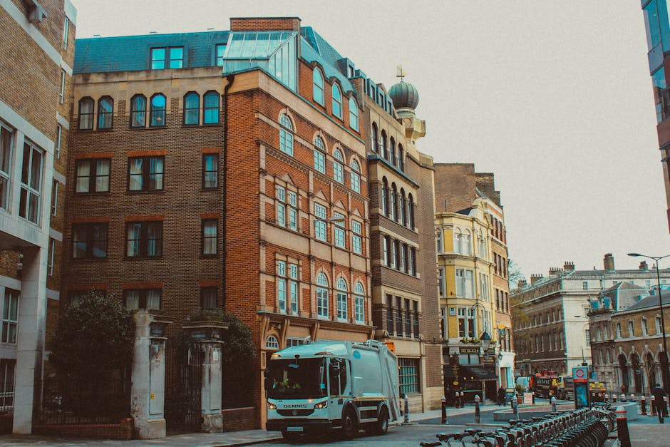 A city street scene featuring a row of historic buildings with intricate facades, large rectangular windows, and decorative architectural elements, situated in an urban environment with a mix of brick