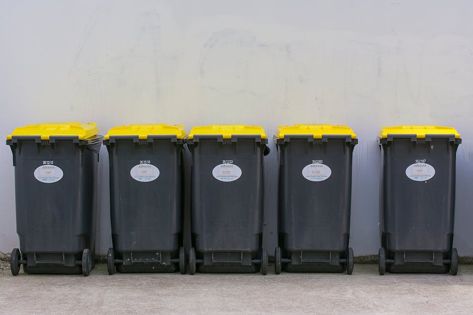In an urban street scene, a large waste collection truck with a blue and white body is positioned near a three-story yellow residential building with balconies featuring black metal railings. The truc