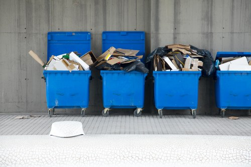 A row of five large, bright blue wheelie bins positioned on a grey pavement outside a concrete wall, filled with mixed waste including cardboard boxes, paper, and plastic packaging. Some bins are partially covered with black plastic rubbish bags, and the contents appear to be overfilled, with waste spilling slightly over the edges. The bins are situated on a textured surface, with a small white object, possibly a piece of paper or cardboard, lying on the ground in front of the bins. The scene reflects typical rubbish collection and waste disposal elements associated with local waste management services in Kentish Town, supporting professional office clearance activities by Office Clearance Kentish Town.