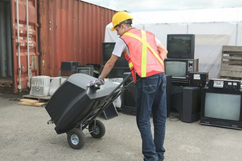 A man wearing a yellow safety helmet, high-visibility vest with pink and yellow reflective stripes, white shirt, and blue jeans is seen pushing a black wheeled rubbish bin toward a collection of discarded electronic appliances. The appliances include old CRT televisions, black stereo speakers, and other electronic waste, all arranged on a paved outdoor surface adjacent to a rust-colored metal fence. In the background, there is a white canopy structure and a wooden pallet, indicating an outdoor waste collection or clearance area. The scene suggests a professional rubbish removal service operating in the Kentish Town area, as part of efforts to clear clutter and manage waste responsibly. The overall lighting is natural, with an overcast sky providing diffuse illumination, emphasizing the practical, work-related context of the image. Office Clearance Kentish Town can assist with rubbish disposal and clearance services involving electrical waste and general rubbish, as visually depicted here.